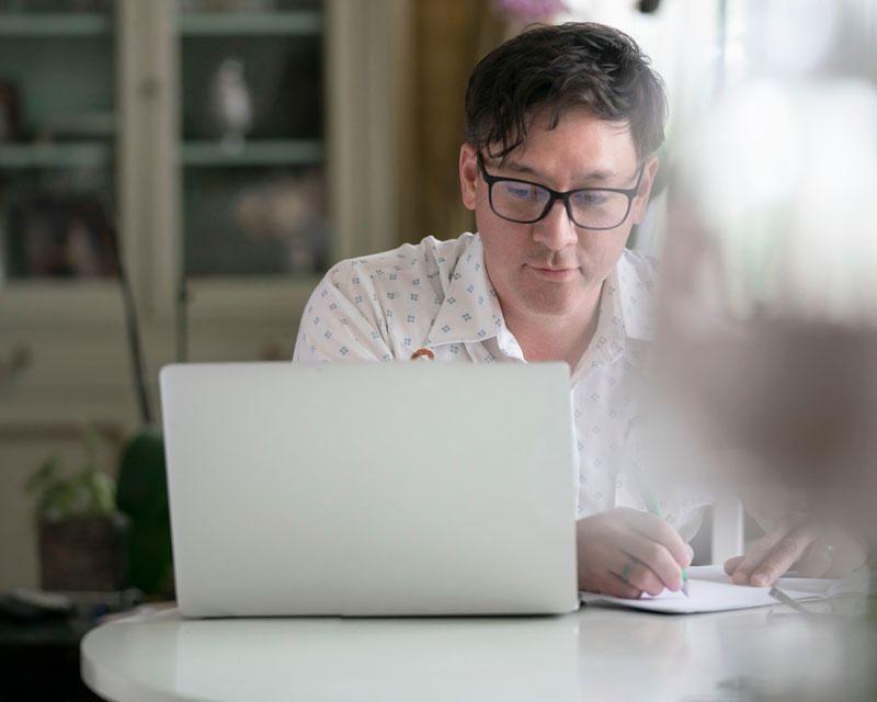Man looks intently at laptop screen while sitting at a home dining room table, takes notes