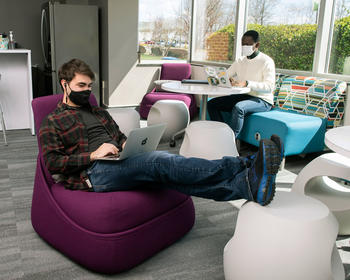 Two graduate students, masked to prevent the spread of coronavirus, work on laptops in the casual atmosphere of the graduate student lounge of the Mason Sci Tech Campus.