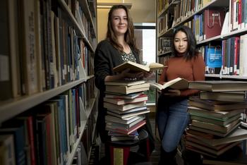 Two women standing in the Mason library stacks with a cart of books and books open in their hands, looking at the camera. 