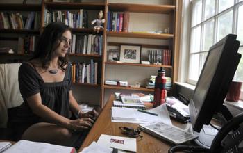 Woman sitting at messy desk looking at desktop computer. Shelves of books in the background.