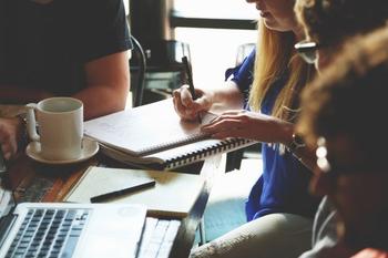 People sitting at table with coffee mugs, pens and notebooks, books, laptops.