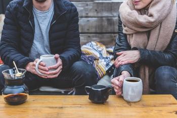 Two people sitting on bench in winter attire with a table and coffee mugs.