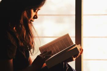 Side profile of woman sitting by a window reading a book.