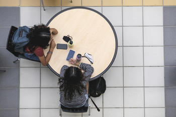 Overhead view of two students, one with a backpack on, sitting at a round table together. Their phones, face masks, and water bottles are on the table.